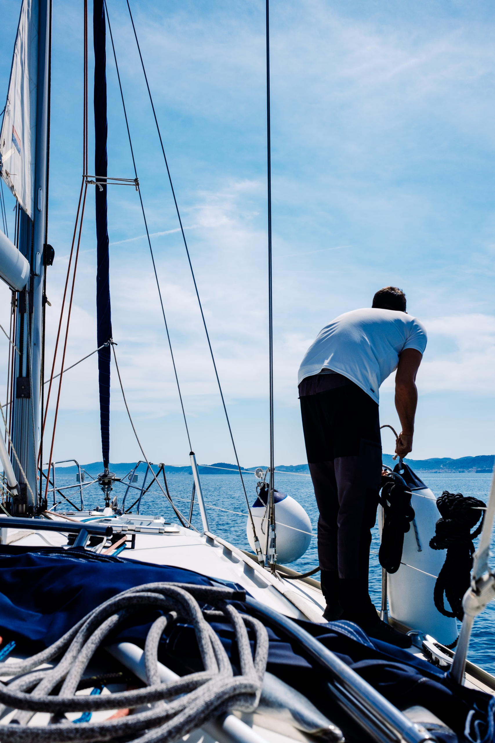 Rear view of a man standing on the deck of a sailboat adjusting boat fenders and ropes while sailing on sunny blue waters.