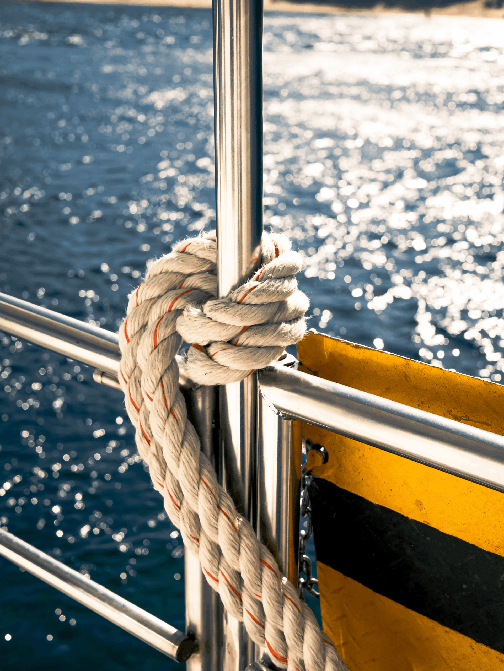 Close-up shot of a thick white marine rope knotted around a stainless steel boat railing over glistening blue sea water.
