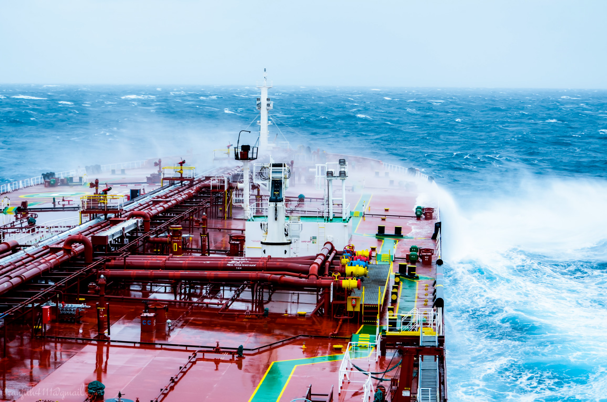 High-angle view of an oil tanker's red deck with industrial pipes navigating through rough, crashing ocean waves.