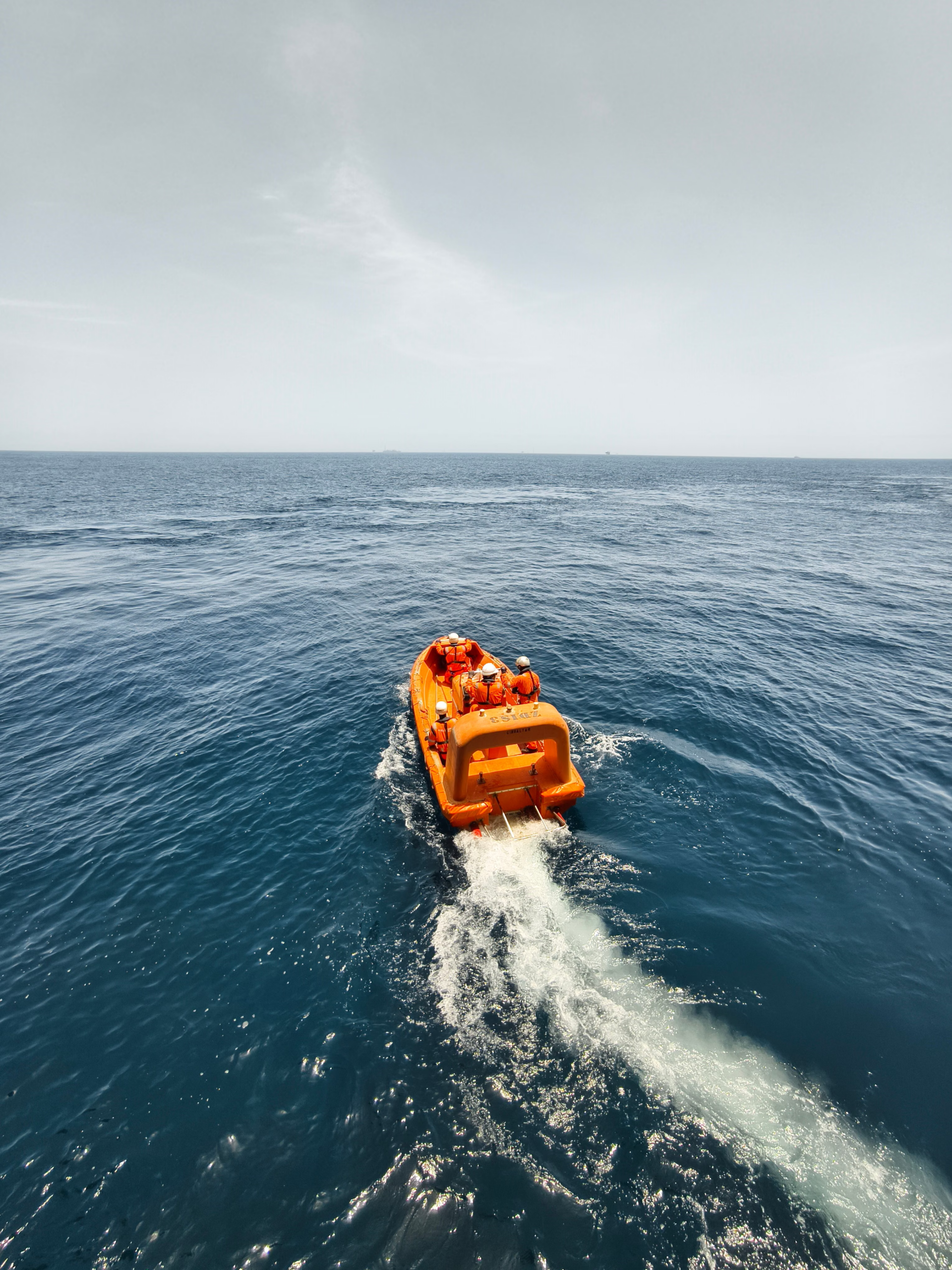 High-angle view of an orange rescue lifeboat with crew members wearing life jackets and helmets, leaving a white wake in the blue sea.