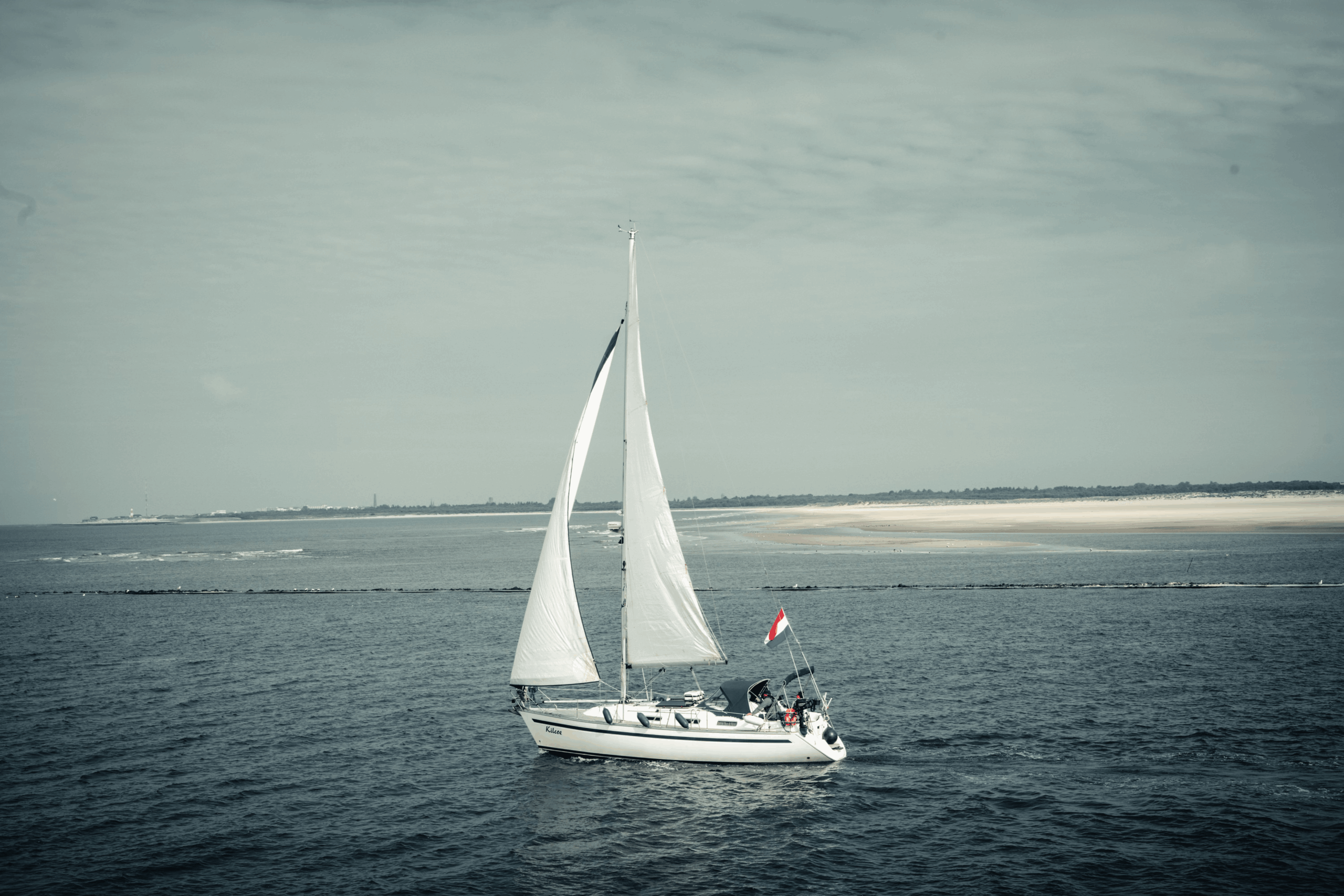 A white sailboat with full sails and a Dutch flag cruising along calm blue ocean waters near a sandy coastline.