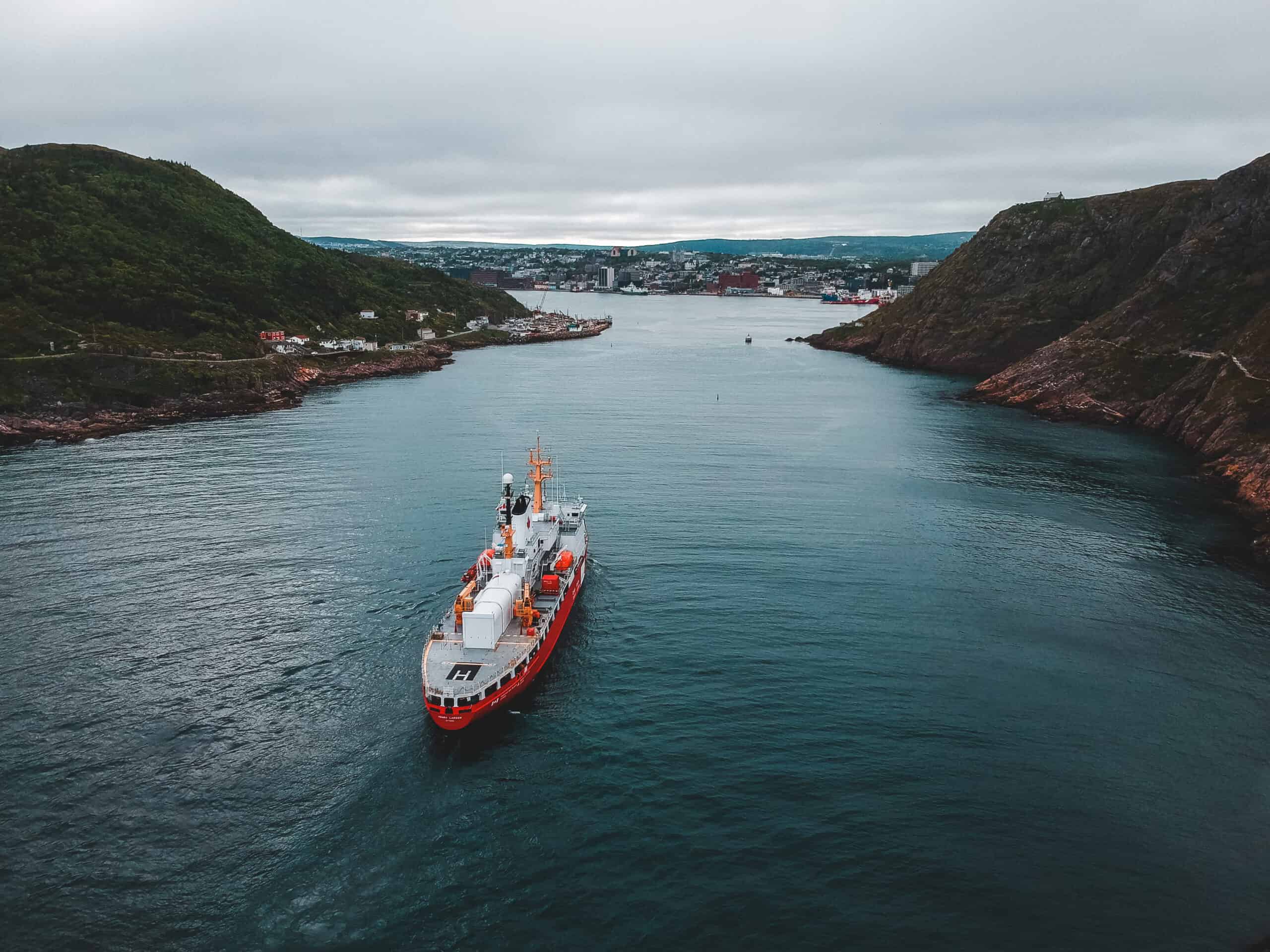 Aerial top-down view of a large red and white vessel sailing through a narrow water channel flanked by steep rocky cliffs and green hills toward a coastal city in the background.