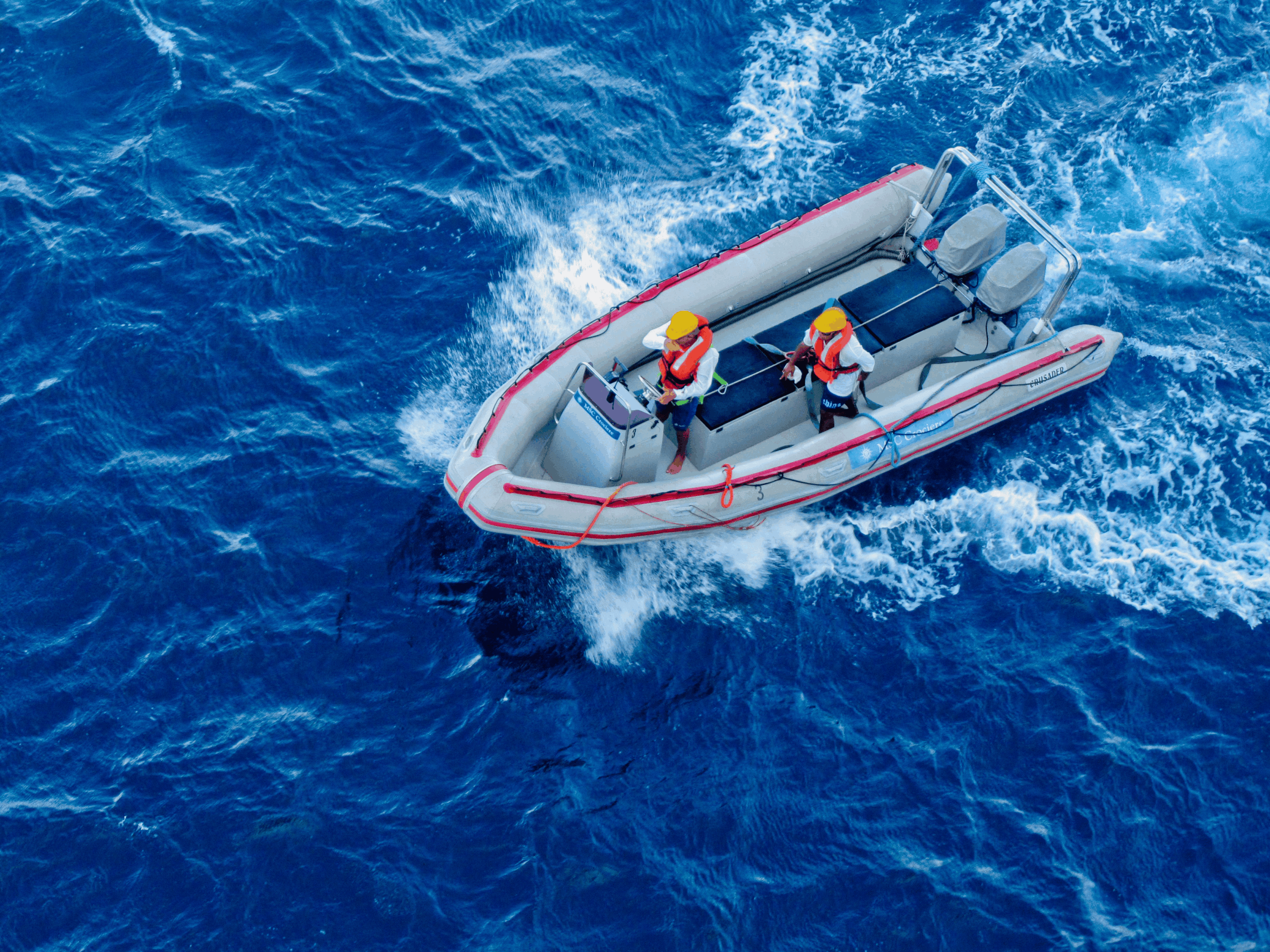 Top-down aerial view of a rigid inflatable boat with two crew members wearing yellow helmets and orange life jackets driving through deep blue water.