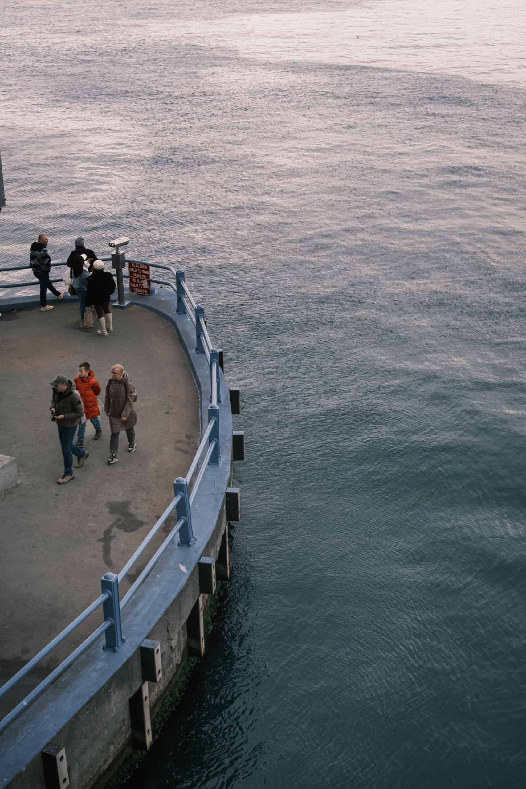 An high-angle aerial view of people walking along a curved concrete waterfront pier with blue railings next to vast open water.